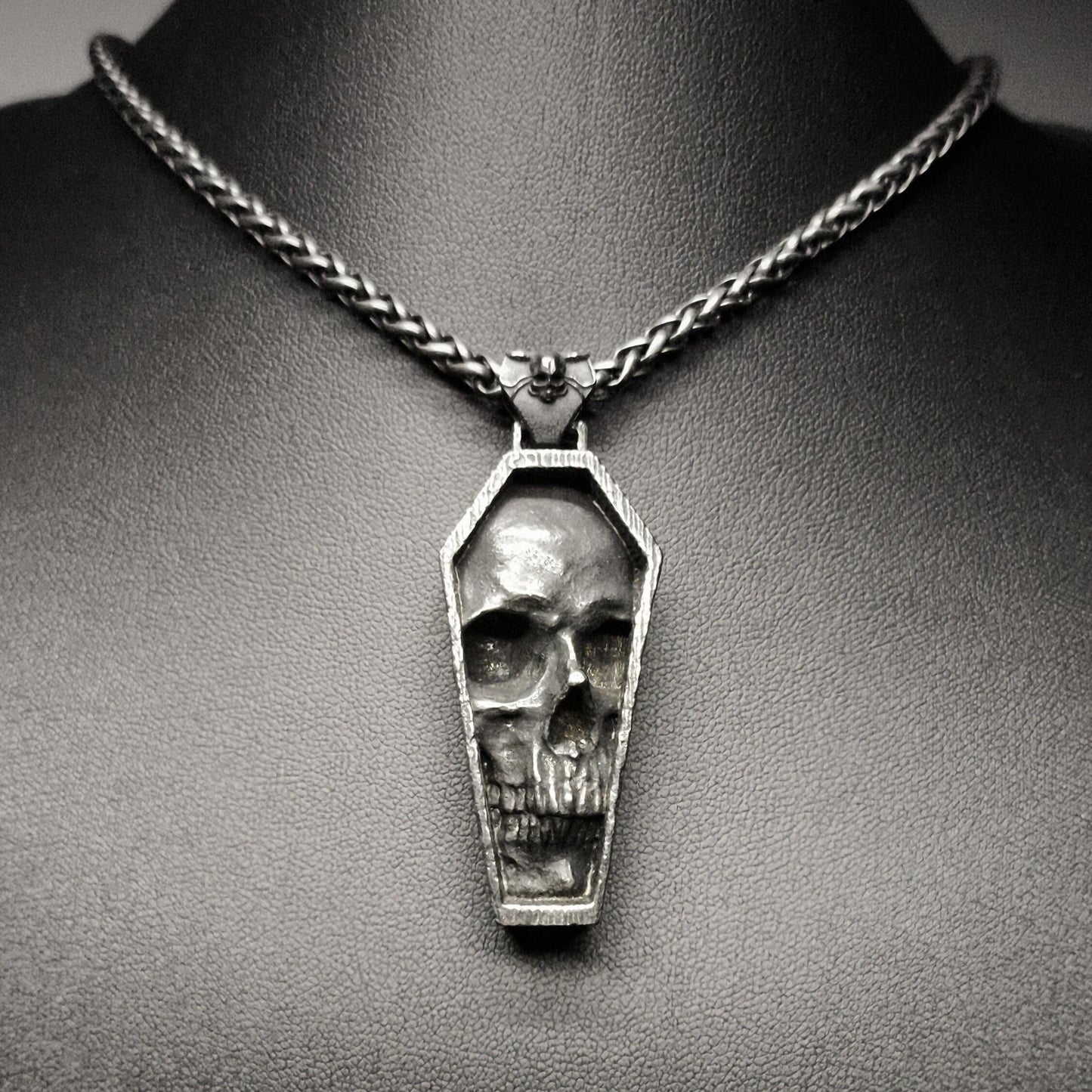 A stainless steel pendant featuring a detailed skull design in a tomb-like structure, displayed on a dark background.