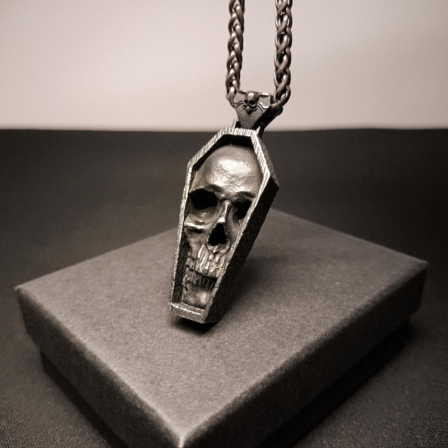 Skull-shaped pendant in a coffin design on a chain, displayed on a gray box.