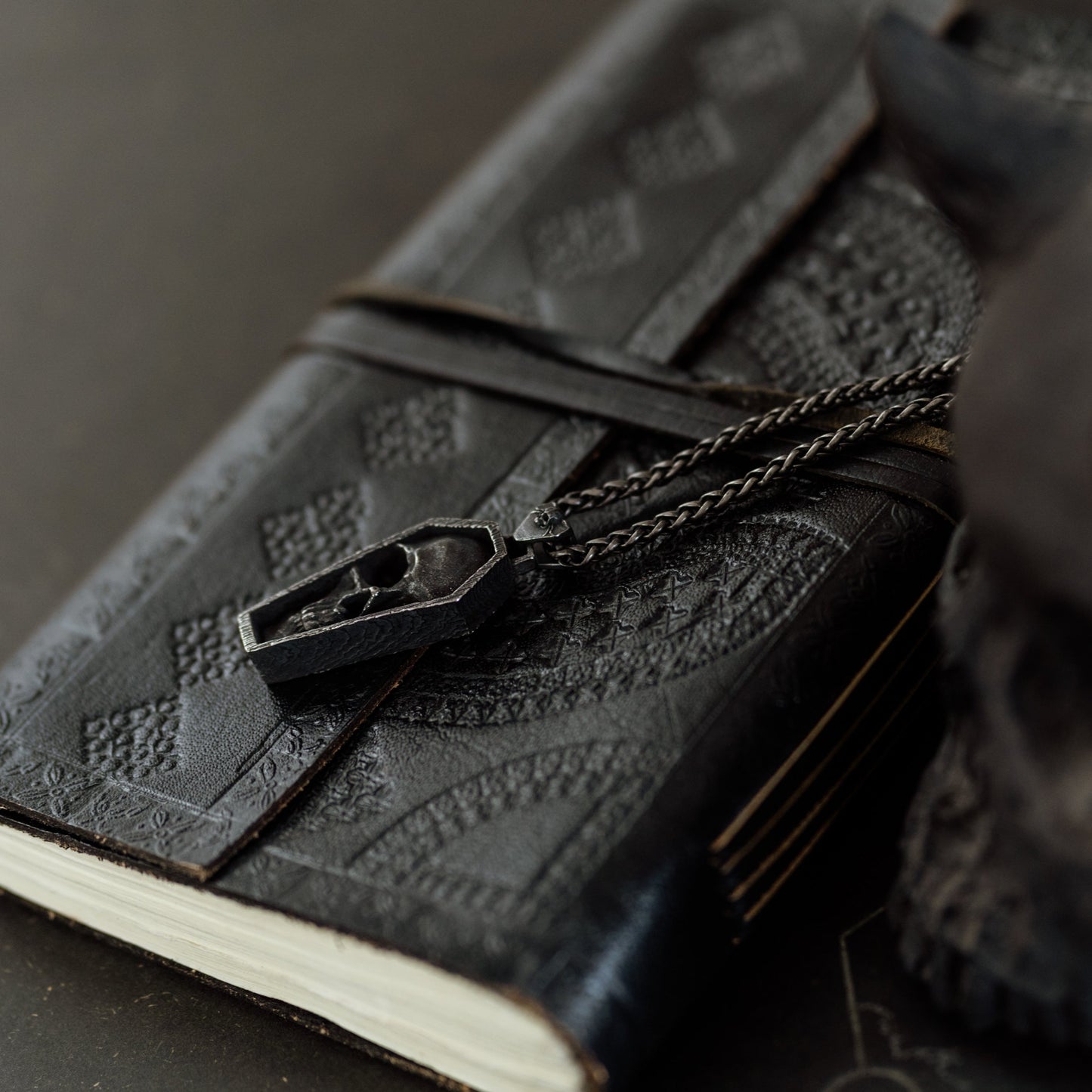 Black leather notebook with embossed design and chain, held by a dark-skinned hand on a dark surface.