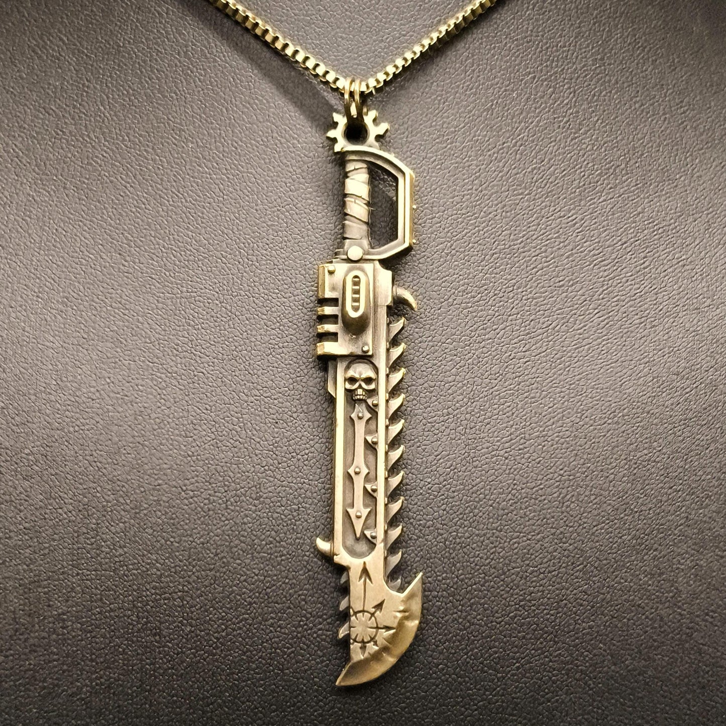 A close-up image of a Warhammer 40K Heretic Chainsword pendant necklace held in a gloved hand, featuring a detailed antique bronze steel pendant with a chain.