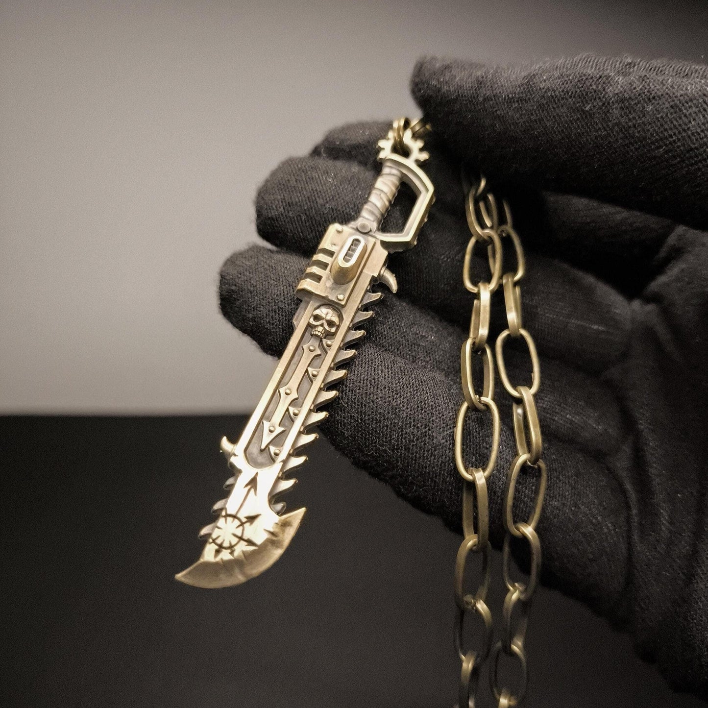 A close-up image of a Warhammer 40K Heretic Chainsword pendant necklace held in a gloved hand, featuring a detailed antique bronze steel pendant with a chain.